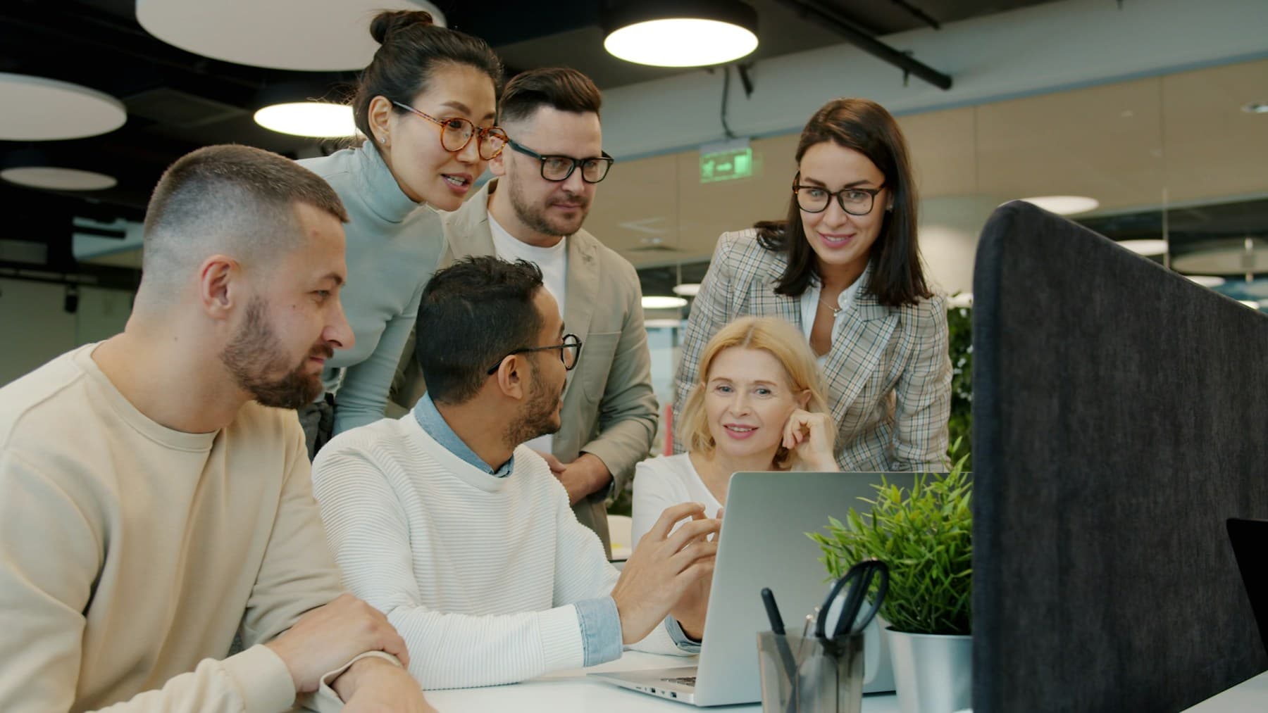 Six colleagues of different ages and backgrounds gathered around a laptop in an open-plan office